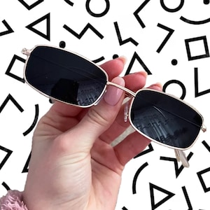 May include: Gold-rimmed sunglasses with black lenses. The sunglasses are held in a hand against a white background with black geometric shapes.