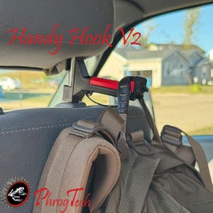 May include: A black and red car headrest hook with the text "Handy Hook V2" and a backpack attached. The backpack is olive green and brown. The logo "Phrog Tech" is in the lower left corner.
