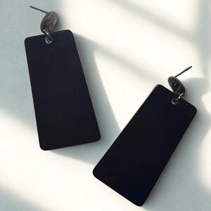 Peut inclure: Une paire de boucles d'oreilles rectangulaires noires avec un clou gris foncé. Les boucles d'oreilles ont un design épuré et minimaliste, avec une forme légèrement effilée. Les boucles d'oreilles sont posées sur un fond blanc.