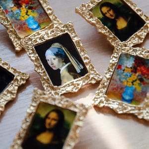 May include: A collection of small, ornate gold picture frames with various paintings inside. The paintings include a portrait of a woman with a blue headscarf, a still life of flowers in a vase, and a portrait of a woman with long dark hair.
