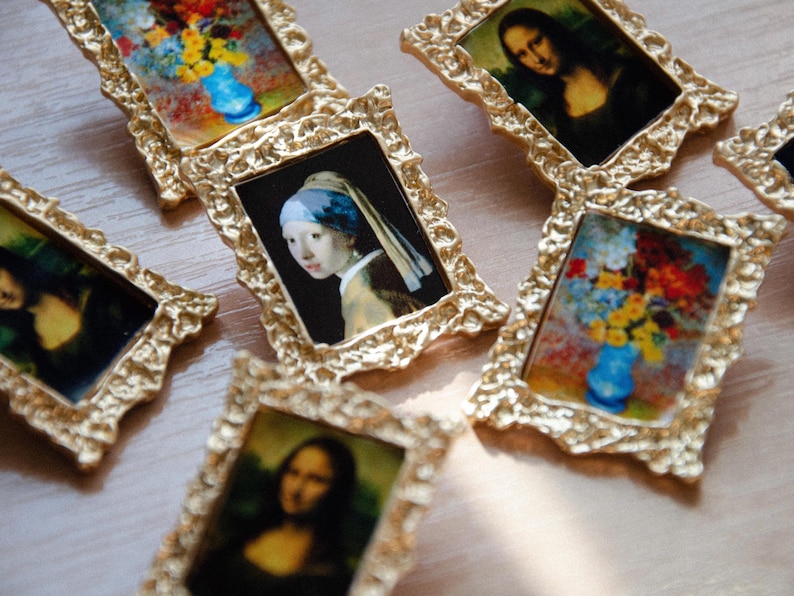 May include: A collection of small, ornate gold picture frames with various paintings inside. The paintings include a portrait of a woman with a blue headscarf, a still life of flowers in a vase, and a portrait of a woman with long dark hair.