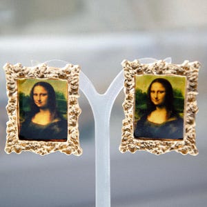 May include: A pair of gold-tone earrings with a textured frame, each featuring a miniature painting of the Mona Lisa.
