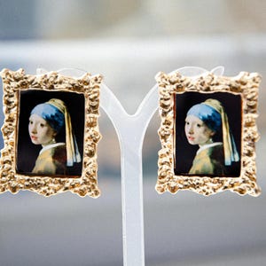 May include: A pair of gold-toned earrings with a miniature painting of the Girl with a Pearl Earring in each. The earrings are on a white display stand.
