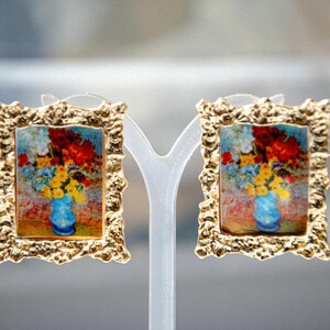 May include: A pair of gold-tone earrings with a textured frame. Each earring features a painting of a vase of flowers in a blue vase.