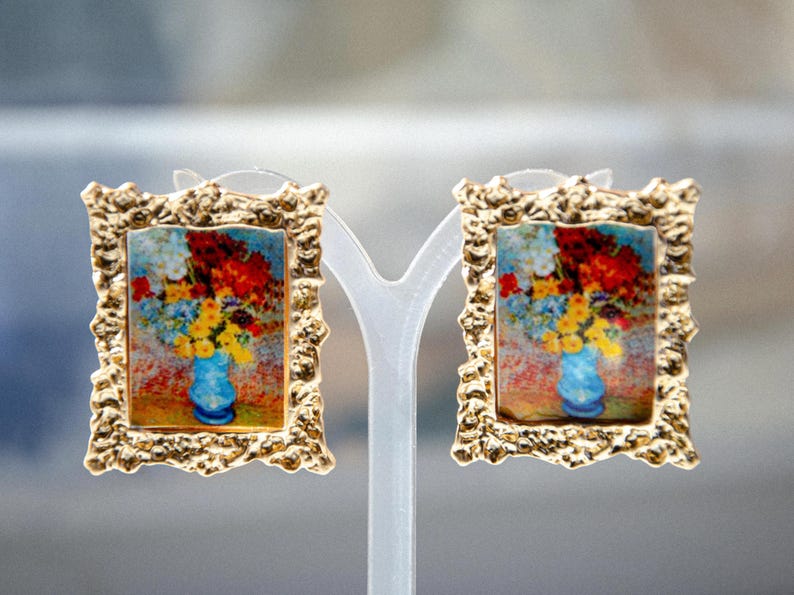 May include: A pair of gold-tone earrings with a textured frame. Each earring features a painting of a vase of flowers in a blue vase.