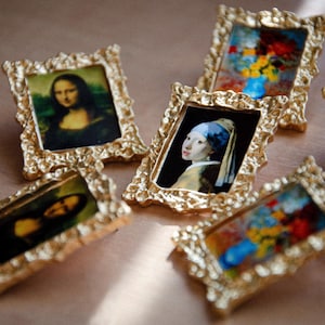 May include: Six small gold-framed pictures with different famous paintings. The pictures are on a light brown surface.