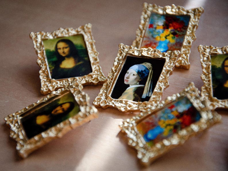 May include: Six small gold-framed pictures with different famous paintings. The pictures are on a light brown surface.