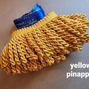 Puede incluir: Primer plano de flecos retorcidos amarillos con una cinta azul. Los flecos están hechos de un material grueso y brillante y tienen una apariencia trenzada. El texto "yellow pinapple" es visible en la imagen.
