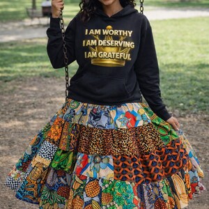 May include: Black hoodie with gold text reading "I AM WORTHY I AM DESERVING I AM GRATEFUL" and a gold crown graphic. The outfit includes a tiered, colorful skirt with various patterns. The skirt is made of multiple fabrics with different designs.