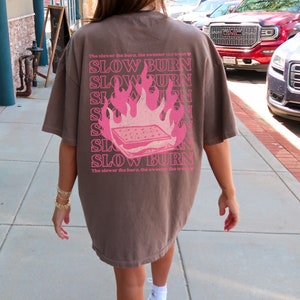 May include: A brown t-shirt with a pink graphic of a burning s'more and the text "The slower the burn, the sweeter the treat" and "SLOW BURN" repeated multiple times.