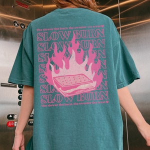 May include: Teal blue t-shirt with a pink graphic of a burning s'more. The text "The slower the burn, the sweeter the treat" is repeated on the shirt.