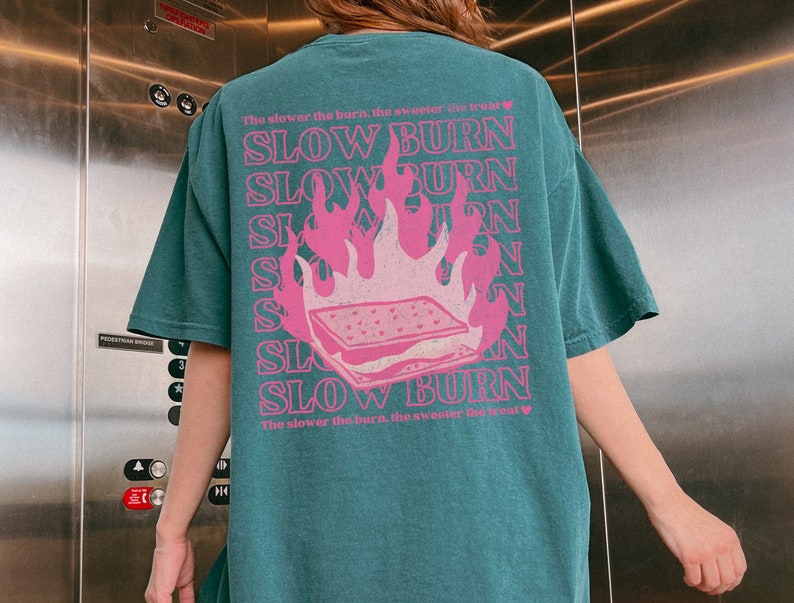 May include: Teal blue t-shirt with a pink graphic of a burning s'more. The text "The slower the burn, the sweeter the treat" is repeated on the shirt.