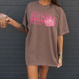 May include: A brown oversized t-shirt with a pink graphic that says "Welcome to Camp Slow Burn" with a campfire and a s'more. The graphic also says "The slower the burn, the sweeter the treat."