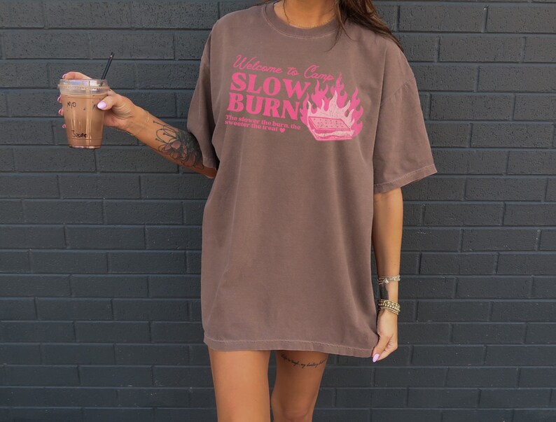 May include: A brown oversized t-shirt with a pink graphic that says "Welcome to Camp Slow Burn" with a campfire and a s'more. The graphic also says "The slower the burn, the sweeter the treat."