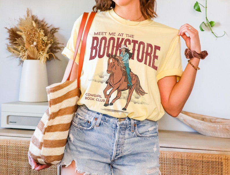 May include: A yellow t-shirt with a graphic of a cowgirl riding a horse. The text on the shirt reads "Meet me at the Bookstore" and "Cowgirl Book Club".