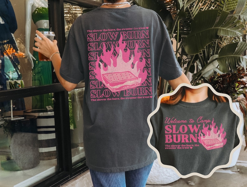 May include: Black t-shirt with a pink graphic of a campfire with a s'more on top. The text "Slow Burn" is repeated multiple times on the shirt. The text "The slower the burn, the sweeter the treat" is also on the shirt.