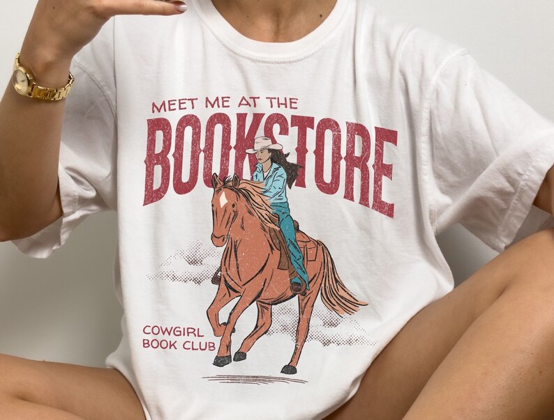 May include: White t-shirt with a graphic of a cowgirl riding a horse. The graphic has the text "Meet me at the Bookstore" and "Cowgirl Book Club".