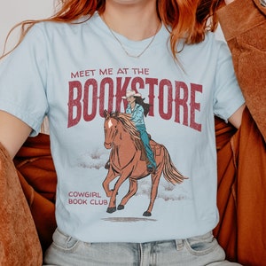 May include: A light blue t-shirt with a vintage graphic design featuring a cowgirl riding a horse. The text on the shirt reads "Meet me at the Bookstore" and "Cowgirl Book Club".