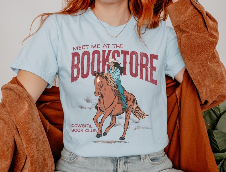 May include: A light blue t-shirt with a vintage graphic design featuring a cowgirl riding a horse. The text on the shirt reads "Meet me at the Bookstore" and "Cowgirl Book Club".