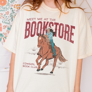 May include: A cream colored t-shirt with a graphic of a cowgirl riding a horse. The text "Meet me at the Bookstore" is printed in red above the graphic. Below the graphic, the text "Cowgirl Book Club" is printed in black.