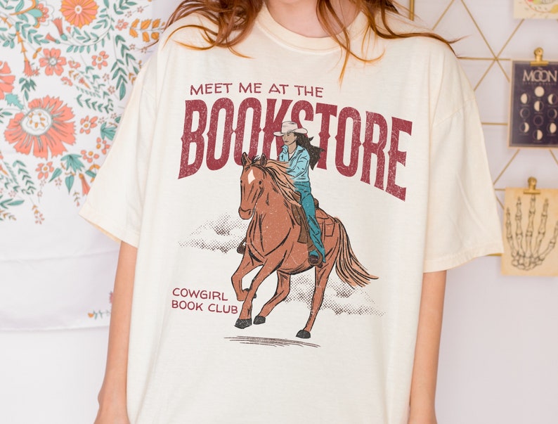 May include: A cream colored t-shirt with a graphic of a cowgirl riding a horse. The text "Meet me at the Bookstore" is printed in red above the graphic. Below the graphic, the text "Cowgirl Book Club" is printed in black.