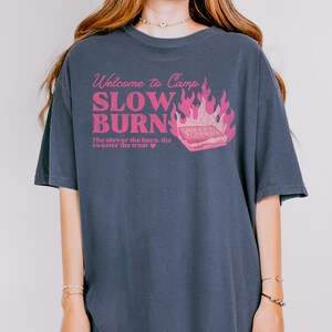 May include: A dark blue t-shirt with a pink graphic that says "Welcome to Camp Slow Burn" with a campfire and a s'more. The text below the graphic says "The slower the burn, the sweeter the treat."