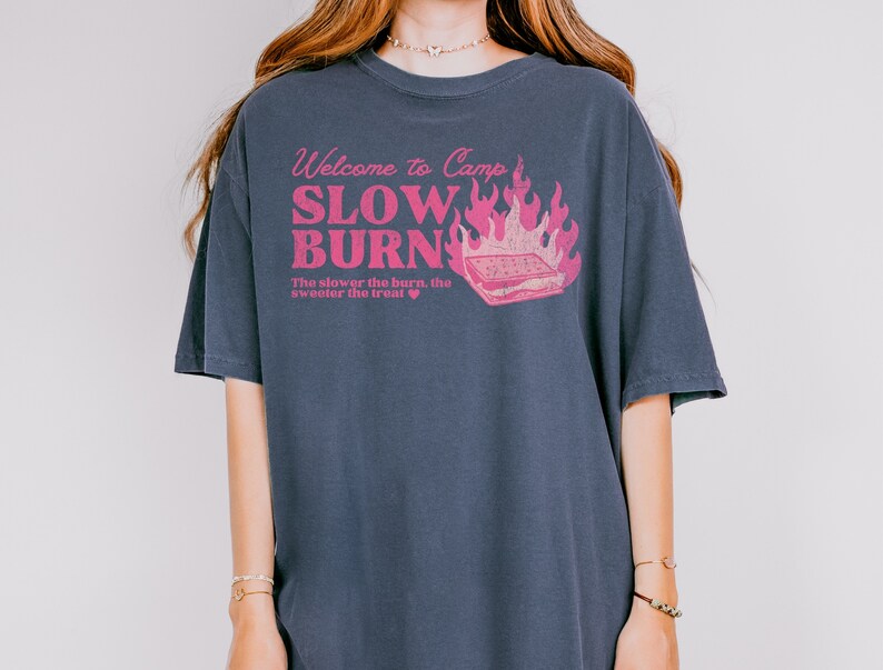 May include: A dark blue t-shirt with a pink graphic that says "Welcome to Camp Slow Burn" with a campfire and a s'more. The text below the graphic says "The slower the burn, the sweeter the treat."