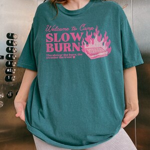May include: Teal blue t-shirt with a pink graphic print that says "Welcome to Camp Slow Burn" with a campfire and a s'more. The text below the graphic says "The slower the burn, the sweeter the treat."