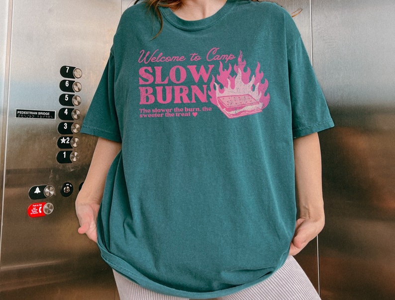 May include: Teal blue t-shirt with a pink graphic print that says "Welcome to Camp Slow Burn" with a campfire and a s'more. The text below the graphic says "The slower the burn, the sweeter the treat."