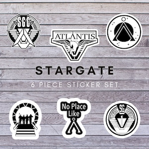 May include: A set of six black and white stickers featuring various symbols and text from the science fiction television series Stargate. The stickers include the Stargate Command logo, the Atlantis expedition logo, the Ancient symbol, the Stargate, the words "No Place Like", and the Stargate Command logo with the number 1.