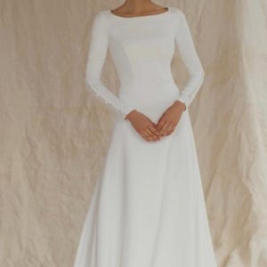 May include: A white wedding dress with long sleeves and a boat neckline. The sleeves have delicate button detailing at the wrists. The dress has a flowing A-line skirt and a subtle train, perfect for a bridal look.