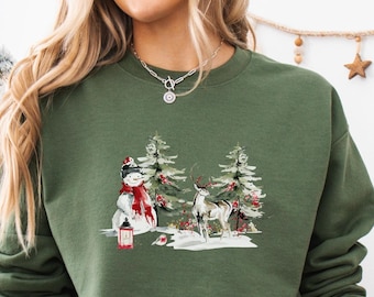 May include: Green sweatshirt featuring a winter scene with a snowman, deer, and pine trees. The design includes a red lantern and snow. The person is wearing light blue jeans and holding a white mug with a Santa Claus graphic.