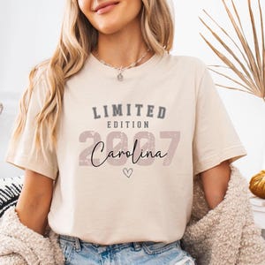 K&ouml;nnte beinhalten: Beiges T-Shirt mit dem Text "LIMITED EDITION 2007 Carolina" in verschiedenen Schriftarten und Farben. Das Shirt wird mit Jeansshorts und einer cremefarbenen Strickjacke kombiniert. Der Look ist l&auml;ssig und entspannt.
