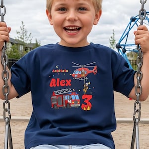Könnte beinhalten: Marineblaues T-Shirt mit rotem und weißem Feuerwehr-Motiv. Das Shirt zeigt einen roten Hubschrauber, einen Feuerwehrwagen, den Namen "Alex" und die Zahl "3". Der Junge lächelt auf einer Schaukel.
