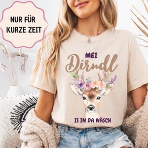 Könnte beinhalten: Beiges T-Shirt mit einem Hirsch-Motiv mit Blumenkranz und dem Text "MEI Dirndl IS IN DA WÄSCH". Eine rosa-weiße Textblase lautet "NUR FÜR KURZE ZEIT". Das Shirt wird mit Jeansshorts getragen.