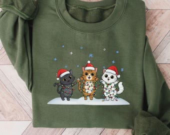May include: Green sweatshirt featuring a festive design of three cartoon cats wearing Santa hats and wrapped in Christmas lights. The cats are standing on a snowy surface with falling snowflakes. The word "Green" is printed at the bottom.