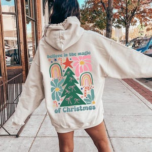 Könnte beinhalten: Sandfarbener Kapuzenpullover mit einem weihnachtlichen Aufdruck auf dem Rücken. Der Aufdruck zeigt einen Weihnachtsbaum, Regenbogen, Blumen und den Text "believe in the magic of Christmas". Der Hoodie ist oversized und hat eine Kapuze.