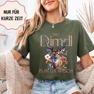 Könnte beinhalten: Olivgrünes T-Shirt mit einer Hirschillustration, floralen Akzenten und dem Text "MEI Dirndl IS IN DA WASCH". Eine rosa-weiße Textblase oben zeigt "NUR FÜR KURZE ZEIT".