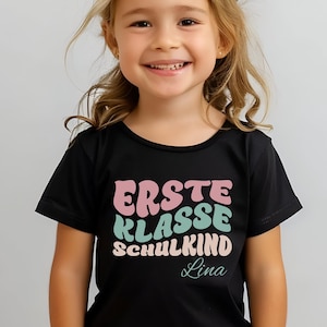 Puede incluir: Una camiseta negra con la frase "ERSTE KLASSE SCHULKIND Lina" en rosa, azul y verde. La camiseta la lleva una niña rubia sonriendo. El texto está en alemán.