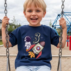 Op de afbeelding: Een marineblauw T-shirt met een cartoon astronaut en een raket met de naam "Anton" erop. Het nummer "3" staat boven de raket gedrukt. De jongen lacht en zit op een schommel.