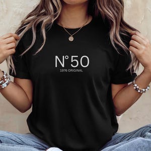 May include: Black t-shirt with the text "N&deg;50 1976 ORIGINAL" in white. The shirt is a classic crew neck style. The model is wearing blue jeans and white sneakers.