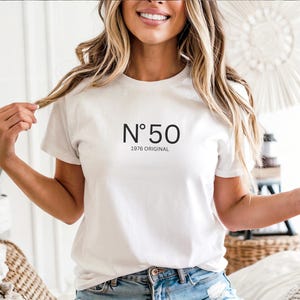 May include: White t-shirt with the text "N&deg;50 1976 ORIGINAL" in black. The shirt is short-sleeved and has a crew neck. The model is wearing blue denim shorts. The shirt is a casual style.