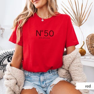 May include: A red t-shirt with the text "N&deg; 50 1976 ORIGINAL" in black. The shirt is short-sleeved and has a crew neck. The model is wearing blue denim shorts and a cream-colored sweater.