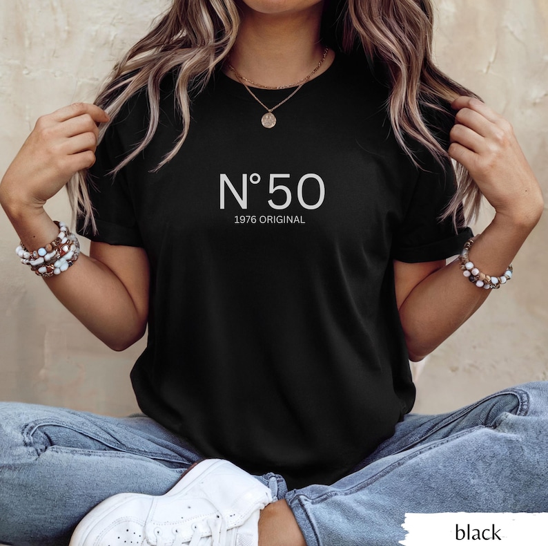 May include: Black t-shirt with the text "N&deg;50 1976 ORIGINAL" in white. The shirt is a classic crew neck style. The model is wearing blue jeans and white sneakers.