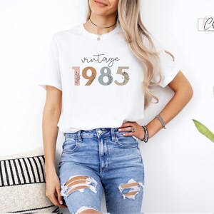 以下が含まれることがあります： 花柄のアクセントが付いたカラフルなフォントで「vintage 1985」と書かれた白いTシャツです。