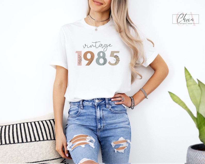 以下が含まれることがあります： 花柄のアクセントが付いたカラフルなフォントで「vintage 1985」と書かれた白いTシャツです。