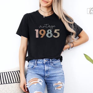 以下が含まれることがあります： カラフルな文字で「ヴィンテージ 1985」と書かれた黒いTシャツ。数字はレトロなスタイルで、花柄のデザインが施されています。