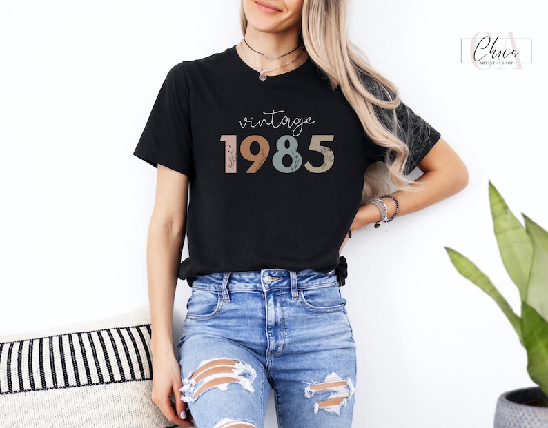 以下が含まれることがあります： カラフルな文字で「ヴィンテージ 1985」と書かれた黒いTシャツ。数字はレトロなスタイルで、花柄のデザインが施されています。