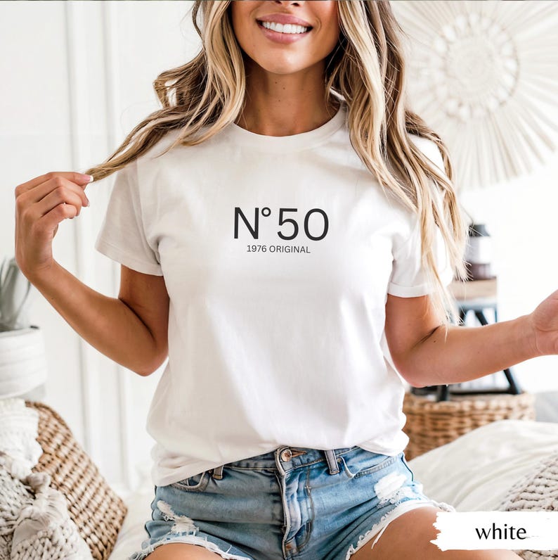 May include: White t-shirt with the text "N&deg;50 1976 ORIGINAL" in black. The shirt is short-sleeved and has a crew neck. The model is wearing blue denim shorts. The shirt is a casual style.