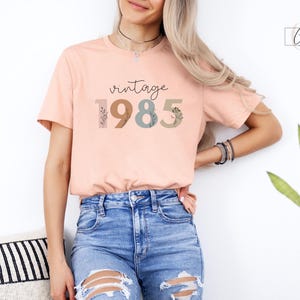 以下が含まれることがあります： 花柄のアクセントがついたスタイリッシュなフォントで「vintage 1985」と書かれた、ピーチ色のTシャツです。